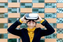 Smiling female in jacket and turtleneck using VR goggles near building windows in daylight — Stock Photo