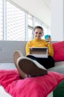 Улыбающаяся взрослая женщина в повседневной одежде и наушниках сидит на диване с подушками и просматривает на планшете с чашкой кофе в светлой квартире — стоковое фото