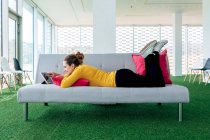 Mulher adulta positiva em roupas casuais deitada no sofá com travesseiro enquanto navega no tablet na sala de estar leve — Fotografia de Stock