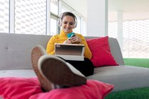 Donna adulta sorridente in abito casual e cuffie seduta su divano con cuscini e navigando su tablet con tazza di caffè in appartamento leggero — Foto stock