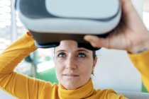 Женщина в повседневной одежде сидит на диване и использует очки VR возле окон в светлом здании — стоковое фото