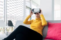 Female in casual outfit sitting on sofa with pillows while using VR goggles near headphones and windows in light building — Stock Photo