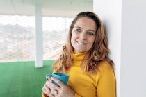 Позитивная взрослая женщина в повседневной одежде с кружкой с кофе в светлой комнате, смотрящая на камеру возле окон и колонн — стоковое фото