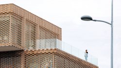 Снизу женщина в стильном наряде стоит на балконе современного здания с геометрическими элементами на окнах при использовании планшета возле стеклянных перил под ярким небом — стоковое фото