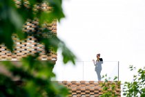 Снизу женщина в стильном наряде стоит на балконе современного здания с геометрическими элементами на окнах при использовании планшета возле стеклянных перил под ярким небом рядом с зелеными деревьями — стоковое фото