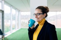 Positive adult female in stylish outfit with mug with coffee in light room looking away near windows and column — Stock Photo