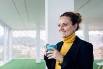 Positive adult female in stylish outfit with mug with coffee in light room looking away near windows and column — Stock Photo