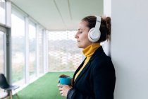 Side view adult female in stylish outfit and headphones with mug with tea with closed eyes leaning on column in light room near windows and green floor — Stock Photo
