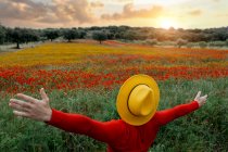 Back view unrecognizable stylish male in red clothes and yellow hat outstretching arms and standing in lush blossoming field in peaceful nature — Stock Photo
