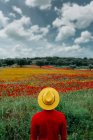 Back view unrecognizable stylish male in red clothes and yellow hat standing in lush blossoming field in peaceful nature — Stock Photo