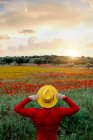 Von hinten unkenntlich stilvoller Mann in roter Kleidung und gelbem Hut, der die Arme ausstreckt und in einem üppig blühenden Feld in friedlicher Natur steht — Stockfoto