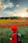 Вид сзади неузнаваемый стильный турист мужчина в красной одежде и желтой шляпе, стоящий в пышном цветущем поле в мирной природе — стоковое фото