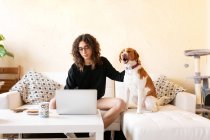 Junge hispanische Hündin streichelt Hund und surft im Internet auf Laptop, während sie ihre Freizeit zusammen im Wohnzimmer verbringt — Stockfoto