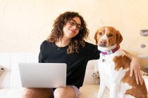 Young Hispanic female stroking dog and browsing internet on laptop while spending free time together in living room — Stock Photo