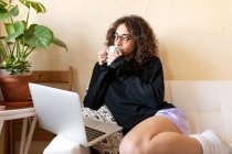 Молодая женщина в повседневной одежде и очках наслаждается горячим кофе и вдумчиво смотрит в сторону, сидя с ноутбуком на диване и охлаждения в одиночестве дома — стоковое фото