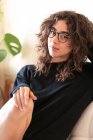 Seitenansicht der jungen lockigen hispanischen Millennial-Frau in heimeliger Kleidung und Brille, die in die Kamera schaut, während sie in der Nähe von Topfpflanzen in einem hellen Raum zu Hause sitzt — Stockfoto