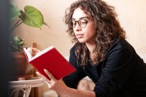 Молода кучерява іспанка в повсякденному одязі і окулярах читає червону книгу і насолоджується цікавою історією під час вільного часу вдома — стокове фото