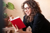 Jeune femme hispanique aux cheveux bouclés heureux en vêtements décontractés et lunettes lire le livre rouge et profiter d'une histoire intéressante pendant le temps libre à la maison — Photo de stock