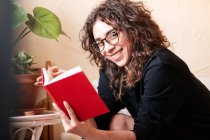 Щаслива кучерява латиноамериканка у звичайному одязі та окулярах, що читає червону книжку та цікаве оповідання під час вільного часу вдома. — стокове фото