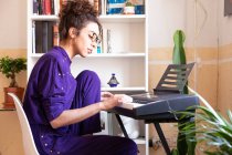 Vue latérale de la jeune femme hispanique jouant du piano électrique tout en pratiquant des compétences musicales à la maison — Photo de stock