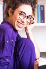 Jeune femme hispanique aux cheveux bouclés en tenue décontractée et lunettes avec des boucles d'oreilles en cerceau penchant la tête sur la main et regardant la caméra tout en se tenant près de la bibliothèque à la maison — Photo de stock