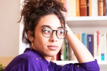 Junge lockige hispanische Frau in Freizeitkleidung und Brille mit Reifrohrringen, den Kopf auf die Hand gestützt und in die Kamera starrend, während sie zu Hause neben dem Bücherregal steht — Stockfoto