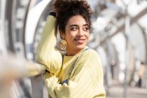 Adolescente hispânica confiante com cabelo encaracolado vestindo macacão jeans e camisola amarela com brincos olhando para longe enquanto se inclina no corrimão na ponte urbana fechada — Fotografia de Stock
