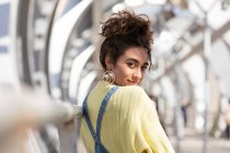 Confident Hispanic female teenager with curly hair wearing denim overalls and yellow sweatshirt with earrings looking at camera while leaning on railing on enclosed urban bridge — Stock Photo