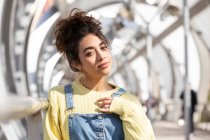 Confident Hispanic female teenager with curly hair wearing denim overalls and yellow sweatshirt with earrings looking at camera while leaning on railing on enclosed urban bridge — Stock Photo