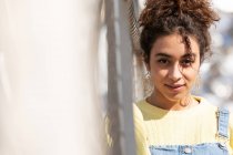Adolescente hispana confiada con pelo rizado que usa overoles de mezclilla y sudadera amarilla con pendientes mirando a la cámara mientras se apoya en la barandilla en un puente urbano cerrado - foto de stock