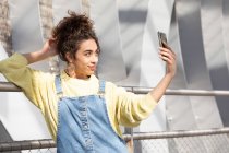 Adolescente hispanique souriante en tenue décontractée tendance et boucles d'oreilles prenant selfie sur smartphone pour partager sur les réseaux sociaux tout en se tenant debout contre un environnement urbain flou — Photo de stock