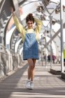 Низкий угол все тело счастливой молодой испаноязычной женщины с вьющимися волосами носить джинсы общее платье с желтой толстовкой и кроссовки ходить по закрытой пешеходный мост в городе — стоковое фото