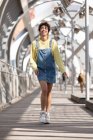 Niedriger Winkel Ganzkörper der glücklichen jungen hispanischen Frau mit lockigem Haar trägt Jeanskleid mit gelbem Sweatshirt und Turnschuhen zu Fuß auf einer geschlossenen Fußgängerbrücke in der Stadt — Stockfoto