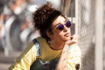 Moderna mujer hispana milenaria con el pelo rizado usando sudadera amarilla con overoles de mezclilla y gafas de sol de moda y pendientes sentados apoyados en la mano cerca de la cerca de malla a la luz del sol - foto de stock