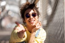 Mulher hispânica milenar moderna com cabelo encaracolado vestindo camisola amarela com macacão jeans e óculos de sol e brincos na moda sentados à mão, chegando à câmera perto da cerca de malha na luz solar — Fotografia de Stock
