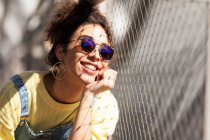 Moderna y alegre mujer hispana milenaria con el pelo rizado usando sudadera amarilla con overoles de mezclilla y gafas de sol de moda y pendientes sentados en la mano cerca de la cerca de malla a la luz del sol - foto de stock
