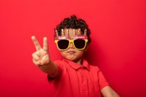 Прохолодна дитина в яскравих сонцезахисних окулярах з днем народження, що показують жест миру на червоному тлі в студії — стокове фото