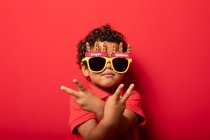Cool enfant portant des lunettes de soleil joyeux anniversaire lumineux montrant geste de paix sur fond rouge en studio — Photo de stock