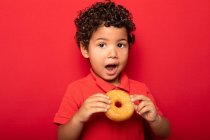 Adorabile bambino con i capelli ricci mangiare dolce ciambella gustosa e guardando la fotocamera su sfondo rosso — Foto stock