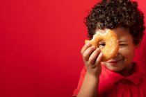 Smiling boy looking at camera through hole in sweet doughnut on red background in studio — Stock Photo