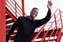 Joven afroamericano encantado con auriculares en el brazo que levanta el cuello en gesto de saludo y mirando a la cámara mientras está de pie en la escalera de metal al aire libre - foto de stock