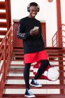 Full body joyful African American male in activewear and headphones browsing mobile phone and standing on metal staircase with gym bag — Stock Photo