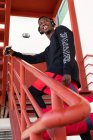 D'en bas joyeux Afro-Américain mâle en vêtements de sport et écouteurs tenant téléphone portable et debout sur un escalier en métal avec sac de gym détournant les yeux — Photo de stock