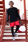 Corps complet joyeux Afro-Américain mâle en vêtements de sport et écouteurs navigation téléphone mobile et debout sur un escalier en métal avec sac de gym regardant la caméra — Photo de stock