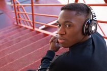 From above content African American male in activewear and wireless headphones sitting on staircase and looking at camera over the shoulder confidently — Stock Photo