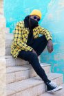 Cool maschio afroamericano in abbigliamento informale alla moda e maschera viso e occhiali da sole e guardando la fotocamera mentre seduto sulle scale in strada — Foto stock