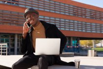 Sonriendo elegante hombre afroamericano con netbook en regazo hablando por teléfono móvil mientras está sentado en la calle soleada - foto de stock
