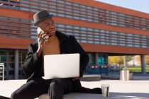Sorridente maschio afroamericano elegante con netbook in grembo parlando sul cellulare mentre seduto su strada soleggiata — Foto stock