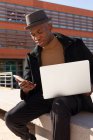 Enfocado hombre afroamericano en ropa elegante y sombrero de navegación teléfono móvil mientras está sentado en la calle con netbook en regazo - foto de stock