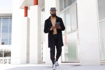 Positiver Afroamerikaner mit niedrigem Winkel in eleganter Kleidung und Gesichtsmaske surft auf dem Tablet, während er auf der sonnigen Straße steht — Stockfoto
